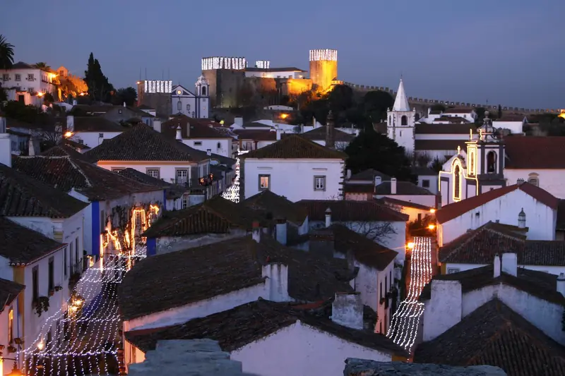 Luzes de Natal nas ruas de Óbidos ao anoitecer, com as torres do castelo iluminadas